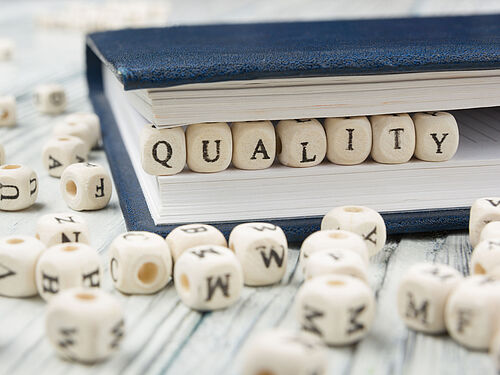 Hölzerne Buchstabenwürfel mit der Aufschrift „QUALITY“ sind teilweise in ein Buch mit blauem Einband gesteckt, umgeben von verstreuten Buchstabenwürfeln auf einer Holzfläche.