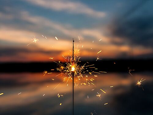 Eine brennende Wunderkerze sprüht helle Funken vor dem Hintergrund eines ruhigen Sonnenuntergangs über einem See, der einen Kontrast zwischen lebendiger Energie und friedlicher Ruhe schafft.