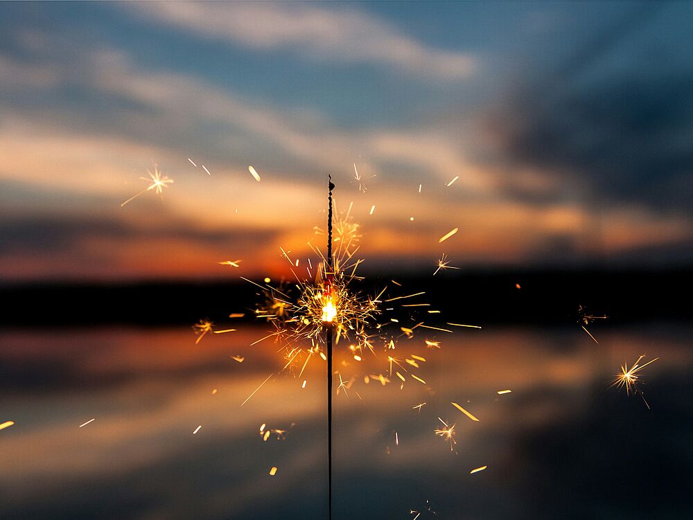 Eine brennende Wunderkerze sprüht helle Funken vor dem Hintergrund eines ruhigen Sonnenuntergangs über einem See, der einen Kontrast zwischen lebendiger Energie und friedlicher Ruhe schafft.