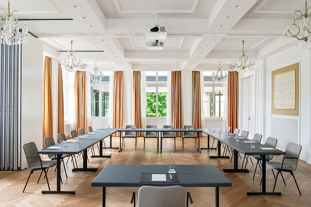 Geräumiger, eleganter Konferenzraum mit großen Fenstern, goldenen Vorhängen, schwarzen Tischen und grauen Stühlen. Die Kronleuchter-Beleuchtung sorgt für eine elegante Atmosphäre.