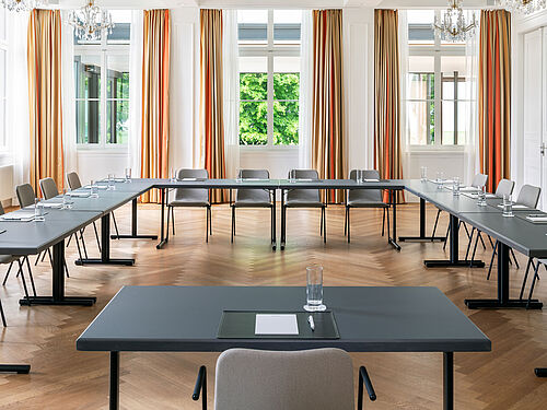 Geräumiger, eleganter Konferenzraum mit großen Fenstern, goldenen Vorhängen, schwarzen Tischen und grauen Stühlen. Die Kronleuchter-Beleuchtung sorgt für eine elegante Atmosphäre.