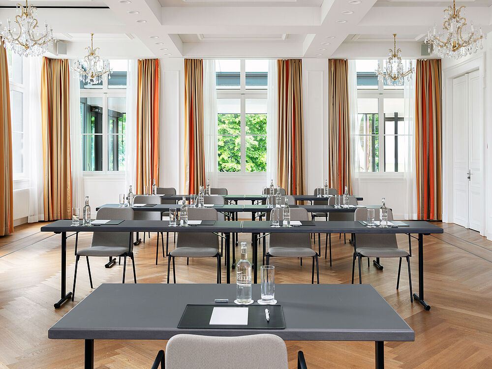 Ein geräumiger, eleganter Konferenzraum mit ordentlich angeordneten Tischen und Stühlen, großen Fenstern mit Vorhängen, Holzfußboden und üppigem Grün im Außenbereich, der einen professionellen Eindruck vermittelt.