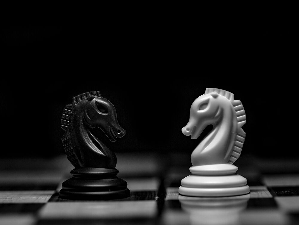 Schwarz-Weiß-Foto von zwei gegnerischen Schachrittern, die sich auf einem Schachbrett gegenüberstehen. Die Szene vermittelt Spannung und Strategie.